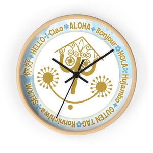 May include: A white clock with a gold stylized tree design and a black clock face. The clock face has a black hour and minute hand. The clock is surrounded by a wooden frame and has greetings in multiple languages around the edge, including "Hello", "Aloha", "Bonjour", "Hola", "Guten Tag", "Konnichiwa", "Ciao", "Selam", "你好", and "Huijambo".