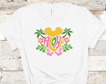May include: White t-shirt featuring a yellow Mickey Mouse head within a pink heart, with the word "Aloha" in a colourful font. The heart is surrounded by green palm leaves and pink flowers.