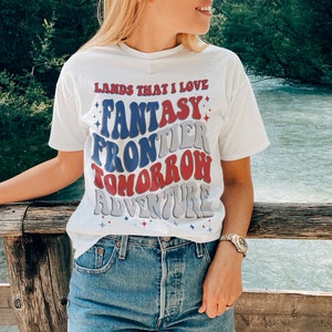 May include: White t-shirt with a retro-style graphic that reads "Lands That I Love Fantasy Frontier Tomorrow Adventure". The graphic is in red, blue, and gray with white stars.
