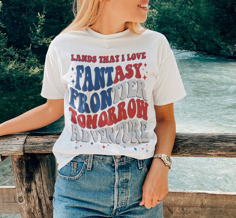 May include: White t-shirt with a retro-style graphic that reads "Lands That I Love Fantasy Frontier Tomorrow Adventure". The graphic is in red, blue, and gray with white stars.