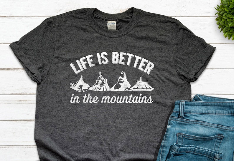 May include: A dark gray t-shirt with the text "Life is better in the mountains" printed in white. The text is surrounded by a white silhouette of a mountain range.