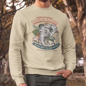 May include: A beige sweatshirt with a graphic print of an elephant and the text "World Famous Jungle Cruise Backside of Water Tours Telling Jokes for Niles and Niles Since July 17, 1955".