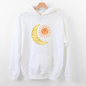 Peut inclure: Sweat-shirt à capuche blanc avec un motif jaune de croissant de lune et de soleil. Le motif comporte le texte "Is there just one moon and one golden sun".