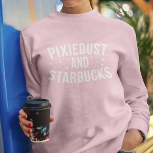 May include: A pink sweatshirt with the text "Pixiedust and Starbucks" in white lettering. The sweatshirt is being worn by a person holding a black coffee cup.