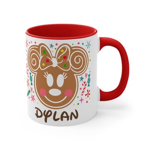 May include: White ceramic mug with a red interior and handle. The mug features a gingerbread Minnie Mouse design with the name "DYLAN" in brown text. Colorful candy and snowflake accents surround the character.