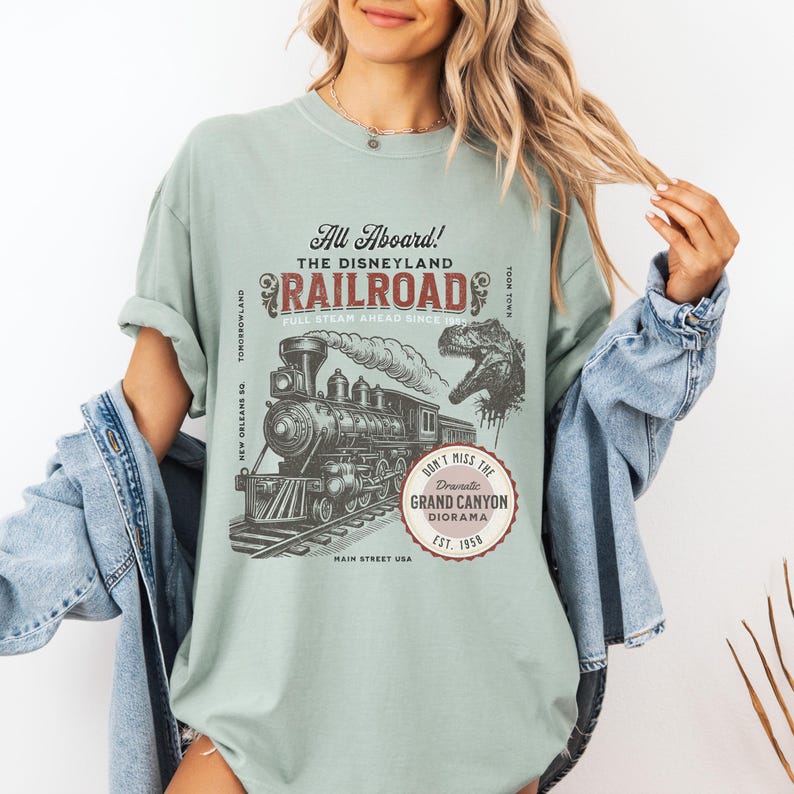 May include: A light green t-shirt featuring a vintage-style graphic of a steam train, the Disneyland Railroad logo, and a dinosaur. The text reads "All Aboard!" and "Don't Miss the Dramatic Grand Canyon Diorama."