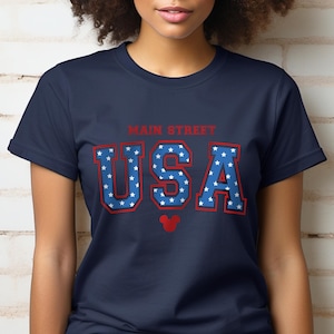 K&ouml;nnte beinhalten: Ein marineblaues T-Shirt mit einer roten und blauen "USA"-Grafik mit wei&szlig;en Sternen. Die Grafik enth&auml;lt auch den Text "Main Street" und einen roten Mickey Mouse Kopf.