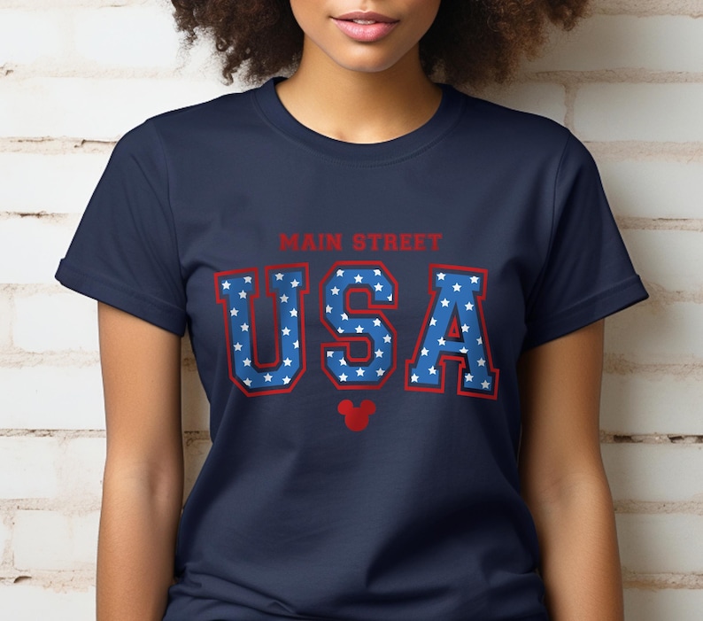 K&ouml;nnte beinhalten: Ein marineblaues T-Shirt mit einer roten und blauen "USA"-Grafik mit wei&szlig;en Sternen. Die Grafik enth&auml;lt auch den Text "Main Street" und einen roten Mickey Mouse Kopf.