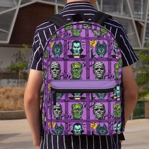 Może przedstawiać: Fioletowy plecak z siatkowym wzorem klasycznych potworów, w tym potwora Frankensteina, mumii i wilkołaka. Plecak ma fioletową przednią kieszeń i czarne paski, idealny do przenoszenia książek i materiałów.
