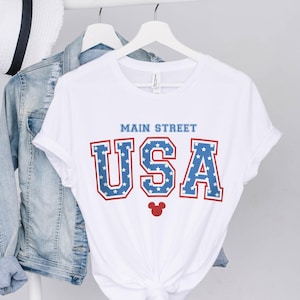 K&ouml;nnte beinhalten: Wei&szlig;es T-Shirt mit einer rot-wei&szlig;-blauen "USA"-Grafik mit Sternen. Der Text "Main Street" befindet sich &uuml;ber der Grafik. Ein roter Mickey-Maus-Kopf befindet sich unter der Grafik.