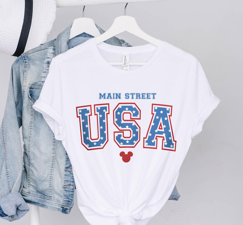 K&ouml;nnte beinhalten: Wei&szlig;es T-Shirt mit einer rot-wei&szlig;-blauen "USA"-Grafik mit Sternen. Der Text "Main Street" befindet sich &uuml;ber der Grafik. Ein roter Mickey-Maus-Kopf befindet sich unter der Grafik.