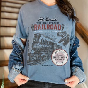 May include: Blue t-shirt featuring a vintage Disneyland Railroad graphic. The design includes a steam train, dinosaur, and text that reads "All Aboard!" and "Grand Canyon Diorama." The shirt is a comfortable, casual style.
