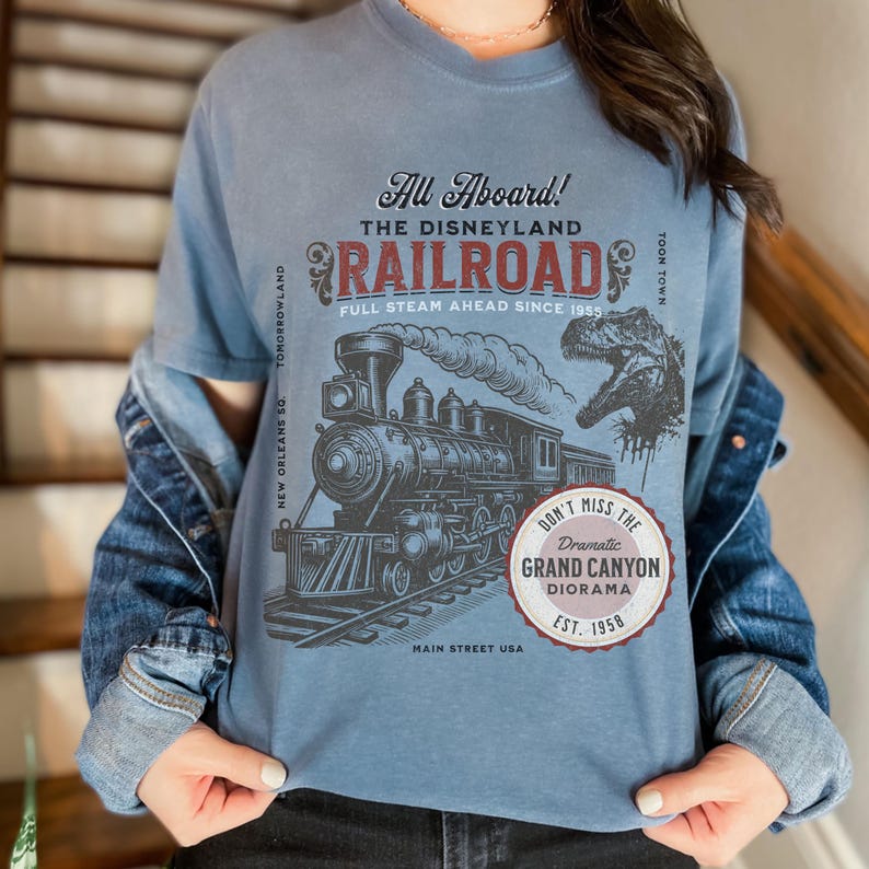 May include: Blue t-shirt featuring a vintage Disneyland Railroad graphic. The design includes a steam train, dinosaur, and text that reads "All Aboard!" and "Grand Canyon Diorama." The shirt is a comfortable, casual style.