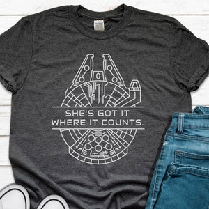May include: Dark gray t-shirt featuring a white line drawing of a spaceship with the text "SHE'S GOT IT WHERE IT COUNTS." The shirt is paired with blue jeans and white sneakers, creating a casual, everyday look. The design is a nod to science fiction.