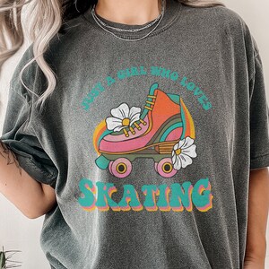 May include: A dark gray t-shirt with a retro graphic design featuring a roller skate with flowers and the text "Just a girl who loves skating".