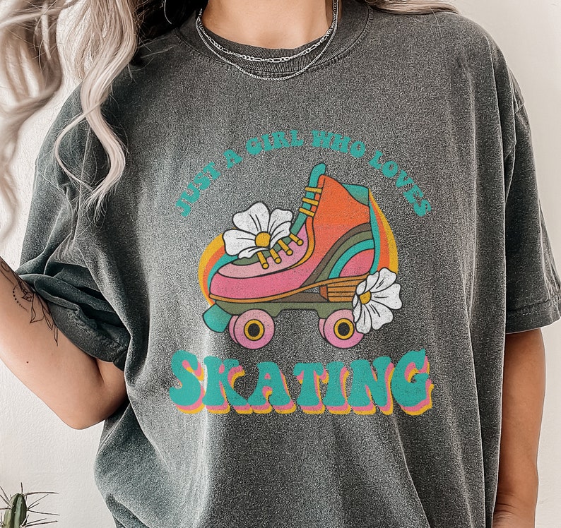 May include: A dark gray t-shirt with a retro graphic design featuring a roller skate with flowers and the text "Just a girl who loves skating".