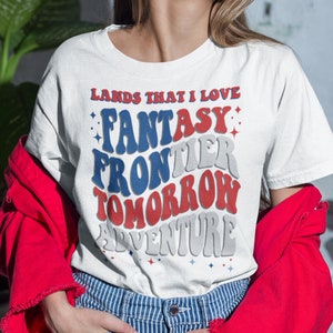 May include: White t-shirt with red, white, and blue text that reads "Lands That I Love: Fantasy Frontier Tomorrow Adventure".