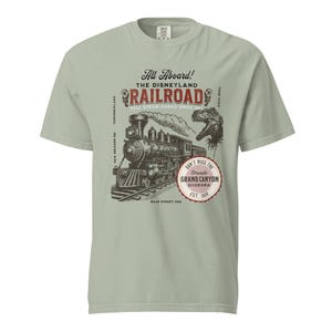 May include: A light green t-shirt featuring a vintage illustration of a steam train on tracks, with the text "All Aboard! The Disneyland Railroad" and "Don't Miss the Grand Canyon Diorama". The design includes a dinosaur and the text "Main Street USA".