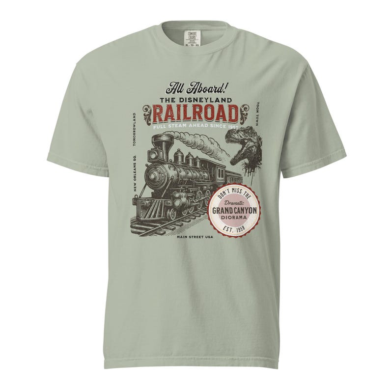 May include: A light green t-shirt featuring a vintage illustration of a steam train on tracks, with the text "All Aboard! The Disneyland Railroad" and "Don't Miss the Grand Canyon Diorama". The design includes a dinosaur and the text "Main Street USA".