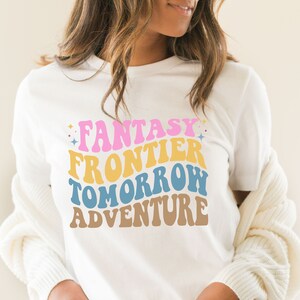 May include: White t-shirt with a retro-style graphic that reads "Fantasy Frontier Tomorrow Adventure" in pink, yellow, blue, and brown.