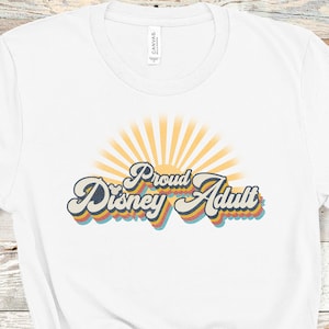 May include: White t-shirt with a retro-style graphic that says "Proud Disney Adult" with a sunburst behind the text.