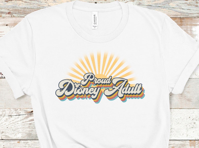 May include: White t-shirt with a retro-style graphic that says "Proud Disney Adult" with a sunburst behind the text.