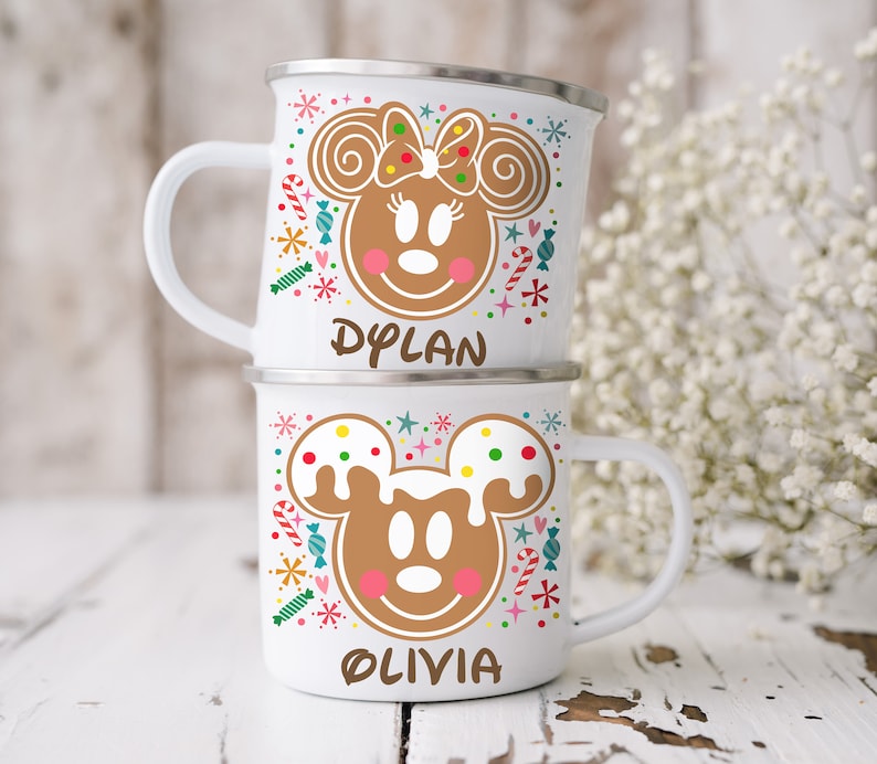 May include: Two white enamel mugs stacked on top of each other. Each mug features a gingerbread-style character design with the names "Dylan" and "Olivia" printed below. The mugs have silver rims and white handles, with colorful candy and star accents.