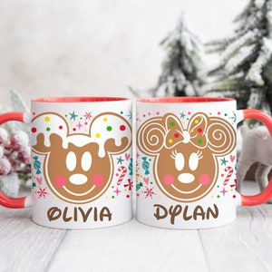 May include: Two white ceramic mugs with red handles and interiors. Each mug features a gingerbread Mickey and Minnie Mouse design with the names "OLIVIA" and "DYLAN" printed below. Festive candy and snowflake accents surround the characters.