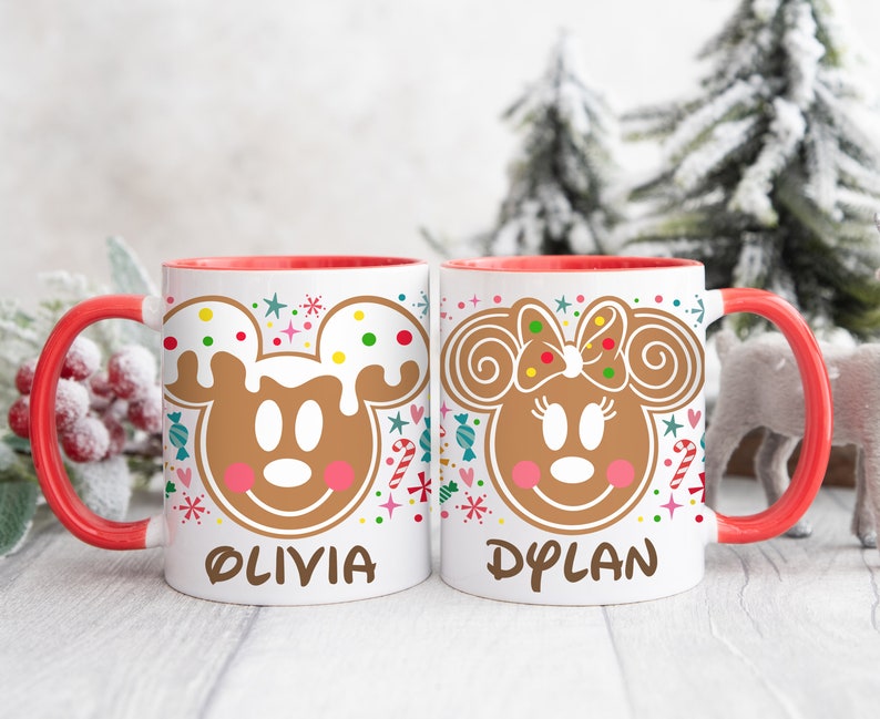 May include: Two white ceramic mugs with red handles and interiors. Each mug features a gingerbread Mickey and Minnie Mouse design with the names "OLIVIA" and "DYLAN" printed below. Festive candy and snowflake accents surround the characters.