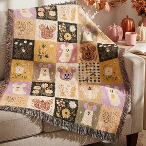 May include: A woven throw blanket featuring a grid pattern of squares with autumn-themed illustrations including ghosts, pumpkins with mouse ears, and floral motifs in shades of orange, yellow, purple, cream, and black with fringed edges.