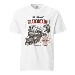 May include: White t-shirt featuring a vintage illustration of a steam train with the text "All Aboard! The Disneyland Railroad." A dinosaur graphic is also included. The design is in black and red.