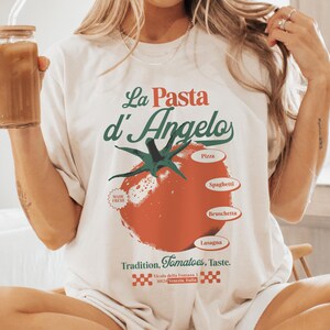 Può includere: T-shirt color crema con la scritta "La Pasta d'Angelo" in rosso e verde. Al centro, una grande grafica di pomodoro rosso, con voci di menu come pizza e spaghetti. La maglietta include anche la scritta "Tradition, Tomatoes, Taste."