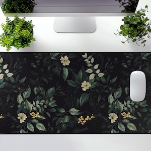 May include: A black desk mat with a floral pattern featuring white flowers and green leaves. The mat has a mouse pad area in the top right corner.