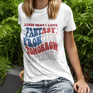 May include: White t-shirt with a red, white, and blue graphic that says "Lands That I Love: Fantasy Frontier Tomorrow Adventure".