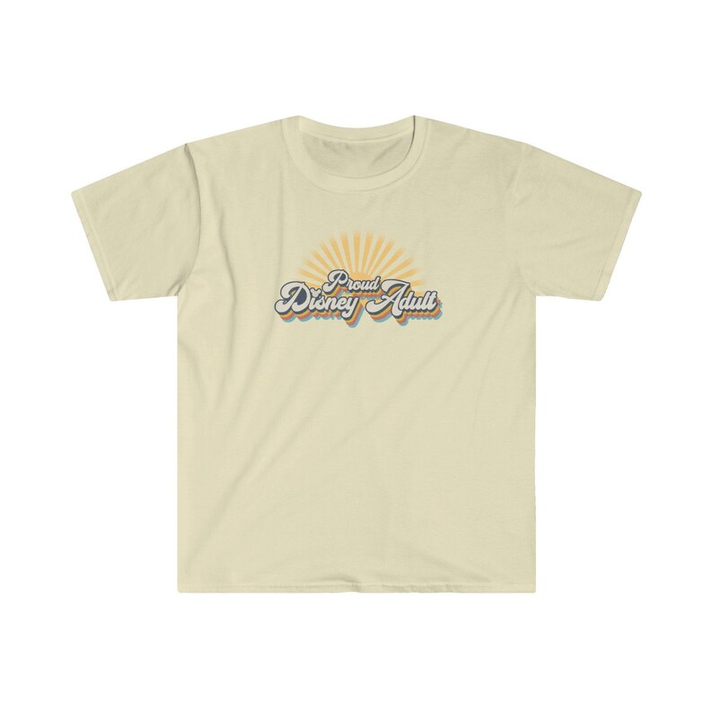 May include: A light beige t-shirt with a retro-style graphic that says "Proud Disney Adult" with a sunburst behind the text. The text is in a blue, orange, and yellow color scheme.