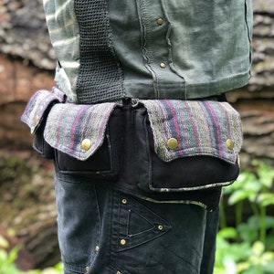 Può includere: Una cintura in tela nera con due grandi tasche e una fodera in tessuto a righe colorate. La cintura è indossata sopra pantaloni neri con più tasche e rivetti dorati.