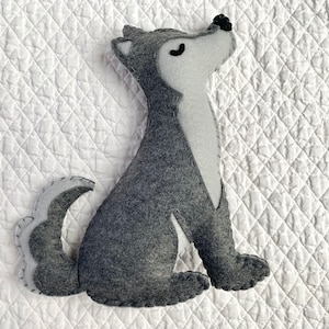May include: A grey felt wolf plush toy with black eyes and a white nose. The wolf is sitting up with its tail curled behind it.