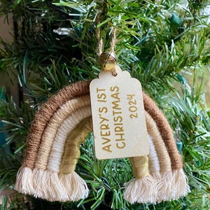 Könnte beinhalten: Ein brauner, beiger und weißer Garn-Regenbogen-Anhänger mit einem Holzschild, auf dem "Avery's 1st Christmas 2024" steht.