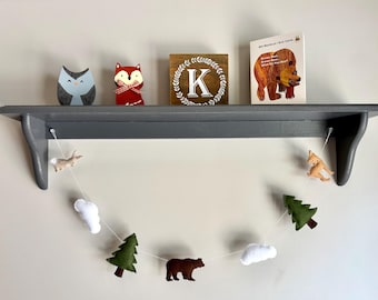 May include: A gray wooden wall shelf holding a blue owl figurine, a red fox figurine with a plaid bow, a square wooden sign with a white letter K, and a Brown Bear, Brown Bear, What Do You See? book. A felt garland with animals, trees, and clouds hangs below.