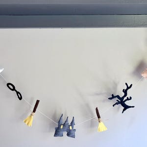 May include: A whimsical garland featuring felt cutouts of a white owl, black glasses, a broom, a castle, a tree branch, a letter, and a lightning bolt. The garland is strung on a white string and is perfect for Halloween or a themed party.
