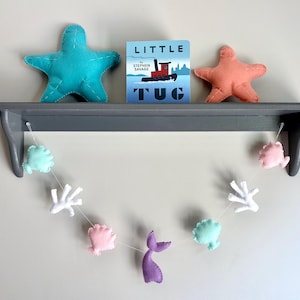 May include: A grey shelf displays a teal starfish, a coral starfish, and a children's book titled "Little Tug." A garland of felt sea creatures, including shells, coral, and a purple mermaid tail, hangs below.