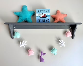 May include: A gray shelf displays a teal starfish, a coral starfish, and a children's book titled "Little Tug." A garland of felt sea creatures, including shells, coral, and a purple mermaid tail, hangs below.