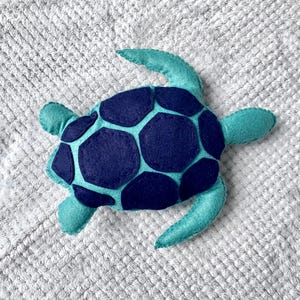 May include: A teal blue felt turtle with a navy blue shell. The turtle is a plush toy with a soft, textured surface.