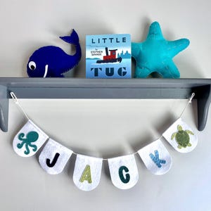 May include: A gray shelf displays a blue whale plush, a children's book titled "Little Tug", and a turquoise starfish plush. Below, a banner spells "JACK" with felt letters and sea creature designs.