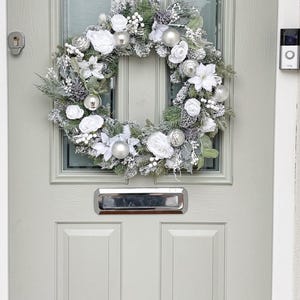 May include: A Christmas wreath with white fabric flowers, silver baubles, and frosted foliage hangs on a light grey door. The door has glass panels and a silver handle. A doorbell is visible.