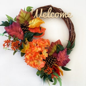 May include: A fall-themed wreath with orange, red, and green leaves, pine cones, and a wooden sign that says "Welcome".