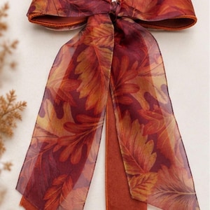 May include: A decorative bow made of sheer, translucent fabric featuring a vibrant print of autumn leaves in shades of deep red, burnt orange, and golden yellow. The bow has two long, flowing tails and a solid rust-coloured fabric backing.