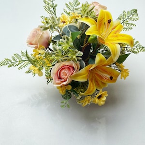 Puede incluir: Un vibrante arreglo floral con lirios amarillos, rosas rosas y pequeñas flores amarillas. El follaje verde y los acentos de helecho complementan las flores. El ramo se coloca sobre un fondo blanco, creando una exhibición fresca y animada.