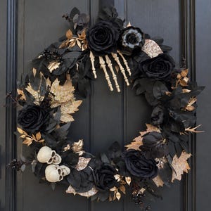 May include: A circular wreath featuring black artificial roses, dark foliage, and gold-toned leaves. The arrangement includes a skeletal hand reaching downward, two small off-white skulls at the bottom, and a dark pumpkin accent.