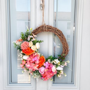 May include: A decorative wreath made of woven twigs, adorned with vibrant faux flowers in shades of pink, orange, and white. The wreath is suspended from a door by a twine cord and a silver spoon.